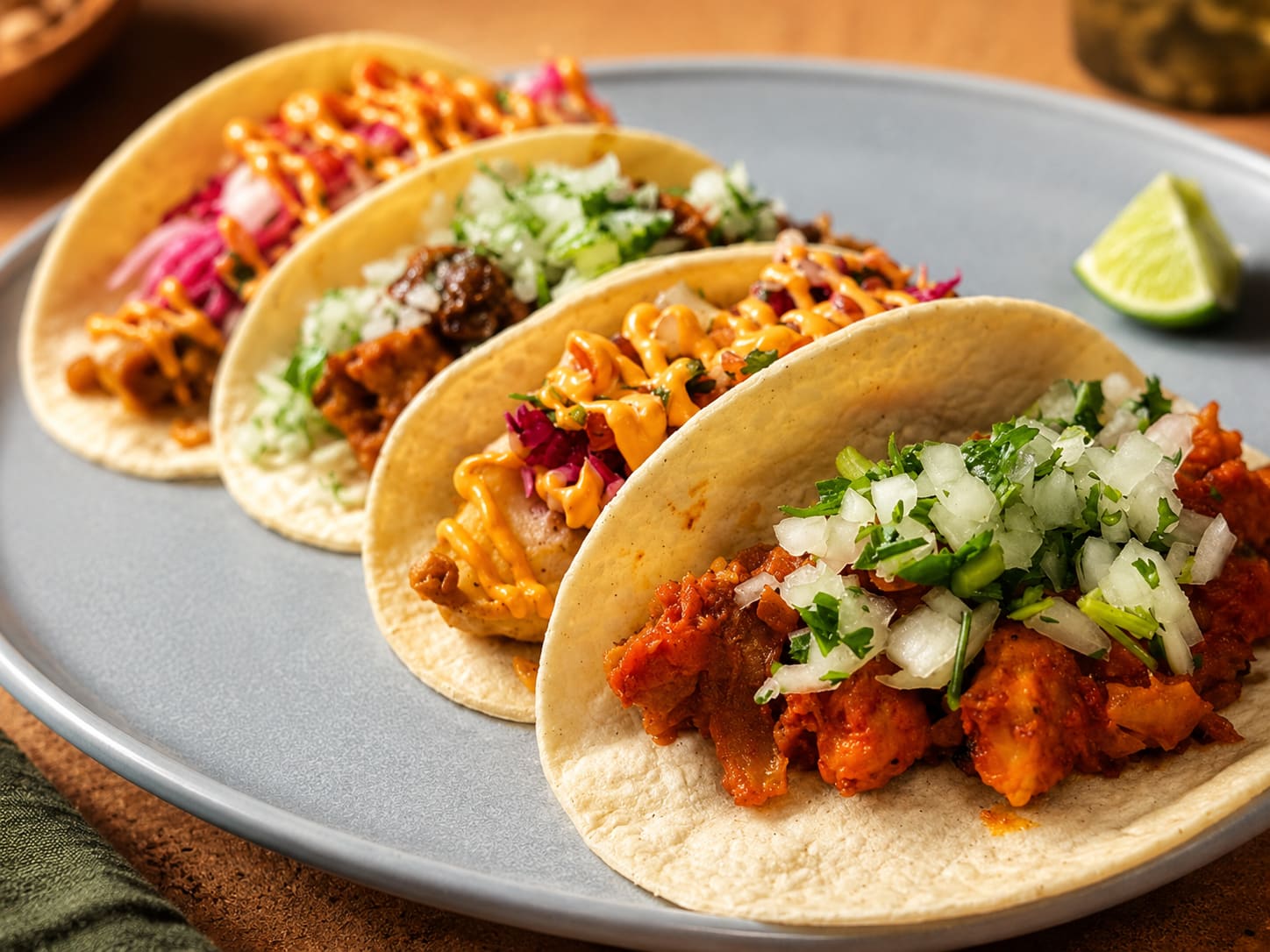 Four tacos on a grey plate — al pastor, carne asada, crispy fish and chicken — with a lime wedge.